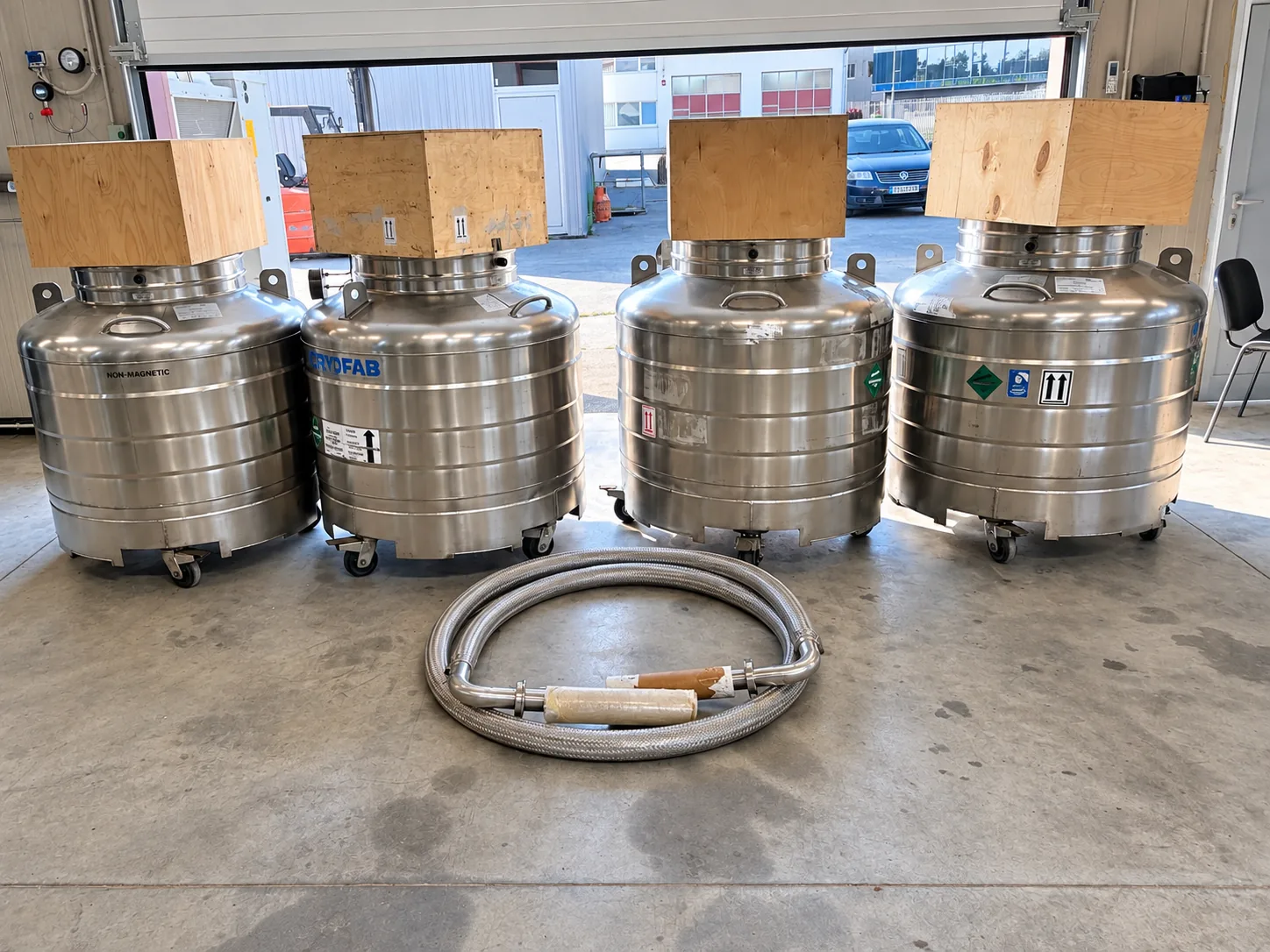 Four stainless-steel cryogenic Dewars with protected upper fittings and a flexible transfer hose